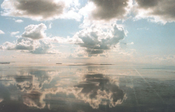 A wide angle shot shows distant islands in a flat mirror-like sea which reflects clouds
