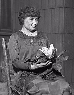 A woman with full dark hair and wearing a long dark dress, her face  in the partial profile, sits in a simple wooden chair. A locket hangs from a slender chain around her neck; in her hands is a magnolia, its large white flower surrounded by dark leaves.